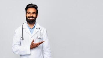 A young Indian man with a beard, wearing a white lab coat and a stethoscope, stands in front of a gray background. He is smiling and gesturing with his hand at copy space photo