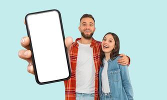 A happy couple is standing together in front of a blue background. The man is holding a black smartphone with a white screen in his right hand. The couple is smiling and looking at the camera. photo