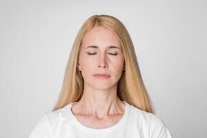A close-up photo of a blonde woman with closed eyes, standing against a simple white background. She appears to be meditating or in a peaceful state.