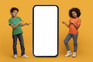 Two happy African American kids are standing on an orange backdrop, each pointing at a large blank screen of a vertically positioned smartphone, conveying a sense of excitement or presentation photo