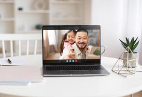 A laptop computer sits on a white table in a well-lit room. The screen displays a call of a man and a young girl, both smiling. photo