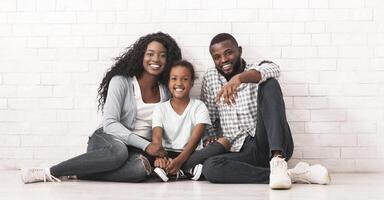 Happy Millennial Family Concept. Smiling Black Parents With Little Daughter Sitting On Floor Together, Smiling And Posing To Camera Over White Brick Wall Background, Free Space photo