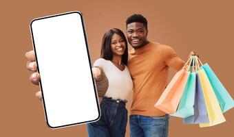 This image shows a happy black young couple holding shopping bags and a smartphone with a blank screen. They are both smiling and looking at the camera. The woman is pointing at the phone. photo