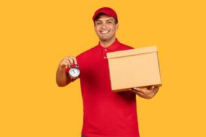 A cheerful delivery man in a red uniform and cap is holding a cardboard box in one hand and an alarm clock in the other. The bright yellow backdrop emphasizes his friendly demeanor and prompt service. photo