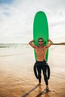 A man in a wetsuit is holding a green surfboard on a sandy beach. He is wearing sunglasses and smiling. The ocean is in the background and the sky is clear. photo