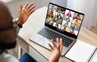 Black man sits at a desk in front of a laptop screen, participating in a conference meeting. He has both hands raised, gesturing to the other participants in the call. photo