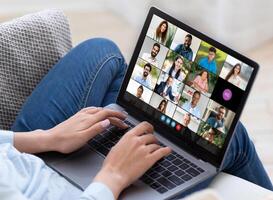 Cropped of woman participates in a virtual team meeting using a laptop while sitting on a couch at home. Several colleagues and friends are visible on the screen during a conference call. photo