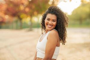 A young woman with curly brown hair is smiling and looking off to the side. She is wearing a white tank top and is standing in a park with trees and grass in the background. photo