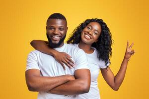 A happy couple poses together against a bright yellow background. The woman has her arm around the man and is making a peace sign with her hand. photo