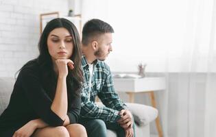 A young woman sits on a sofa with a troubled expression, her chin resting on her hand. She wears a black top and appears to be upset. A man sits next to her, looking away with a serious expression. photo