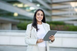 A young businesswoman, dressed in a white blazer and a white top, smiles confidently as she holds a tablet computer in front of a modern building. The scene portrays a professional setting photo