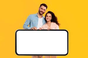 A young man and woman are holding a large blank smartphone in front of them, both smiling at the camera. The background is a solid yellow color. photo