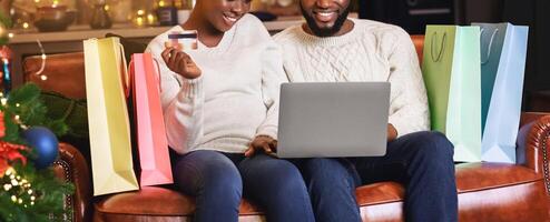 Christmass shopping. Smiling black couple buying Xmas presents online, using laptop and credit card while sitting on sofa in living room, panorama photo