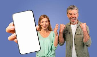 This image features a couple, a man and a woman, excitedly holding a smartphone with a blank white screen. They are both smiling and looking at the camera. The background is a solid blue color. photo