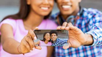 A teen black couple is taking a selfie with a smartphone while smiling. Guy is holding the phone in front of them, while the girl is smiling and looking at the phones screen. photo