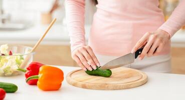 A woman is shown cutting a cucumber on a wooden cutting board. She is using a sharp knife to slice the cucumber into smaller pieces. The cucumber is green and fresh, cropped photo