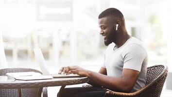 A smiling man works on his laptop while sitting at a table in a cafe. He is wearing earbuds and has a focused expression. photo