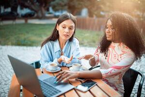 Two women are seated at a table outside a cafe. One woman is using a laptop computer while the other woman gestures and looks on. There is a cell phone on the table next to the laptop. photo