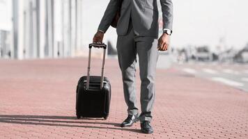Unrecognizable black businessman walking with rolling suitcase outside a modern building, ready for a business trip or travel. photo