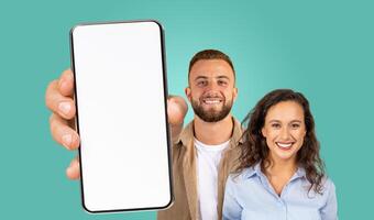 A man and a woman are standing together against a teal background. The man is holding a smartphone with a blank screen towards the camera. They are both smiling and looking directly at the camera. photo