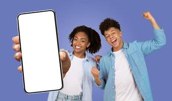Black couple stand in front of a purple background. The woman on the left holds a smartphone in front of her, pointing at the screen with her index finger, smiling with her teeth showing photo