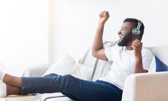 African American man wearing headphones is sitting on a couch and listening to music. He is smiling and has his hand raised in the air, as if he is dancing or singing along to the music. photo