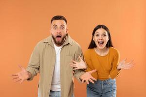 A young couple stands together, both displaying expressions of surprise and excitement with wide eyes and open mouths. They are dressed casually and the background is a solid orange photo