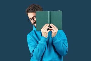 A young man with glasses smiles mischievously while holding a green book in front of face. He wears a blue hoodie and stands against a solid backdrop, creating a lighthearted and vibrant atmosphere. photo