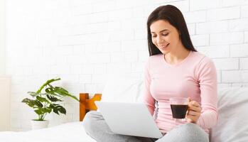 A woman is seated on a bed holding a cup of coffee in one hand and attentively looking at a laptop screen. She appears focused and engaged in the content displayed on the laptop, blank space photo