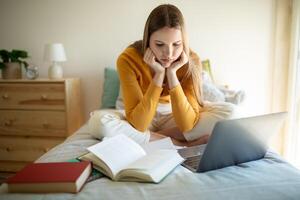 A teen girl sits on her bed in a cozy, well-lit bedroom. She is surrounded by open books and using a laptop, deeply focused on her work or studies. photo