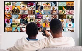 Two men are sitting on a couch and watching television. The television screen is showing a mosaic of various images, depicting different scenes and activities. photo