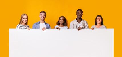 Smiling group of multiracial students standing together in row and displaying white empty placard, yellow studio background photo