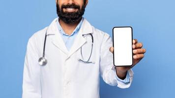 A doctor wearing a white coat and stethoscope smiles while holding a smartphone with a blank screen towards the camera. He is standing in front of a blue background. photo