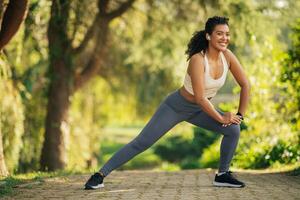 Woman wearing athletic clothes stretching in a park. She is standing on a paved path with green trees and grass in the background. She smiling and appears to be enjoying her workout. photo