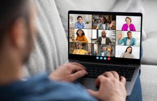A man sits in a casual setting with a laptop on his lap, actively participating in a conference with multiple participants. The screen displays a diverse group of people photo