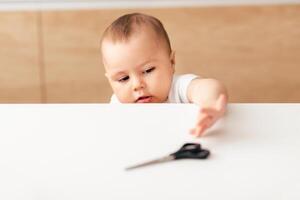 A baby is reaching out with curiosity towards a pair of sharp scissors on a table, posing a potential danger to the child. photo