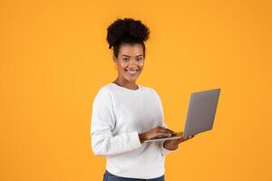 A young woman with a bright smile uses a laptop while standing in front of a yellow background. She is dressed in a white sweater and appears to be working or browsing the internet. photo