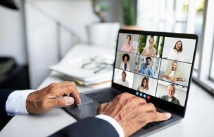 Unrecognizable man is sitting in front of a laptop, engaging in a call with a group of people displayed on the screen. He appears focused and attentive as he interacts with his virtual audience. photo