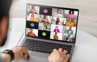 Businessman is using a laptop computer to participate in a conference meeting with multiple colleagues. The laptop screen displays a grid of feeds, showing the faces of each participant photo