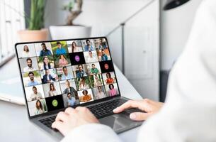 A woman is seated in front of a laptop, engaging with a group of people visible on the screen. She appears focused and attentive, possibly in a virtual meeting or online conference, cropped photo