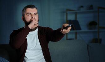 A bearded man in a dark sweater sits on a couch and yawns while holding a TV remote. The living room is dimly lit, creating a cozy atmosphere with shelves and household items in the background. photo