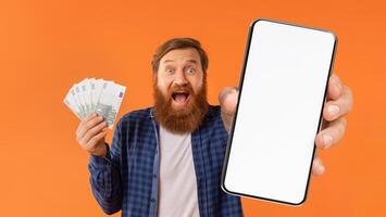 A cheerful man holds a handful of cash in one hand and a large smartphone in the other. His joyful expression and the bright orange backdrop create an energetic atmosphere. photo