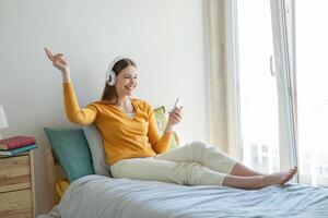 A girl is lounging on her bed, enjoying music through her headphones. She is smiling, relaxed, and appears to be in a light, airy room with natural sunlight streaming in through a large window. photo
