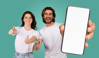 A man and a woman stand in front of a green background, smiling and giving thumbs up. The man is holding a smartphone with a blank white screen, facing the camera. Both are wearing white t-shirts. photo