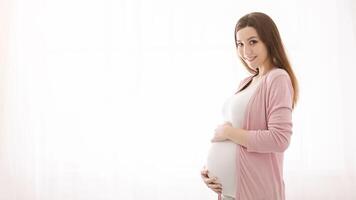 A pregnant woman is smiling while gently holding her belly. She appears joyful and content as she embraces her baby bump, copy space photo