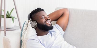 Black man with a beard is sitting on a couch, leaning back with a relaxed expression. He is wearing headphones and listening to music. He has a big smile on his face photo