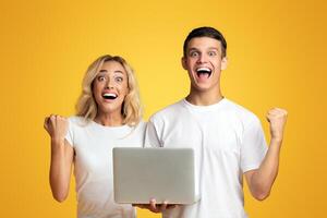 Happy millennial guy and girl celebrating win with laptop, shouting on yellow studio background, panorama with copy space photo