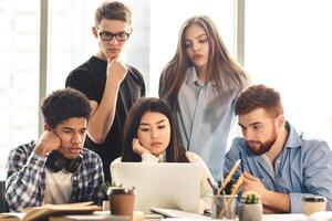 A group of five young professionals is gathered around a laptop, collaborating intently on a project. Some appearing pensive and others focused, they work together in a sunlit office setting photo