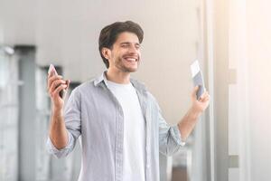 Enjoying Travels. Portrait Of Happy Young Man Standing In Airport, Holding Passport With Tickets And Smartphone In Hands, Raising Hands In Excitement photo