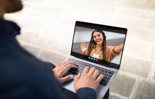 A man is seated outdoors, using a laptop to chat with a woman. He is typing on the keyboard while she smiles and waves at him from the screen. photo